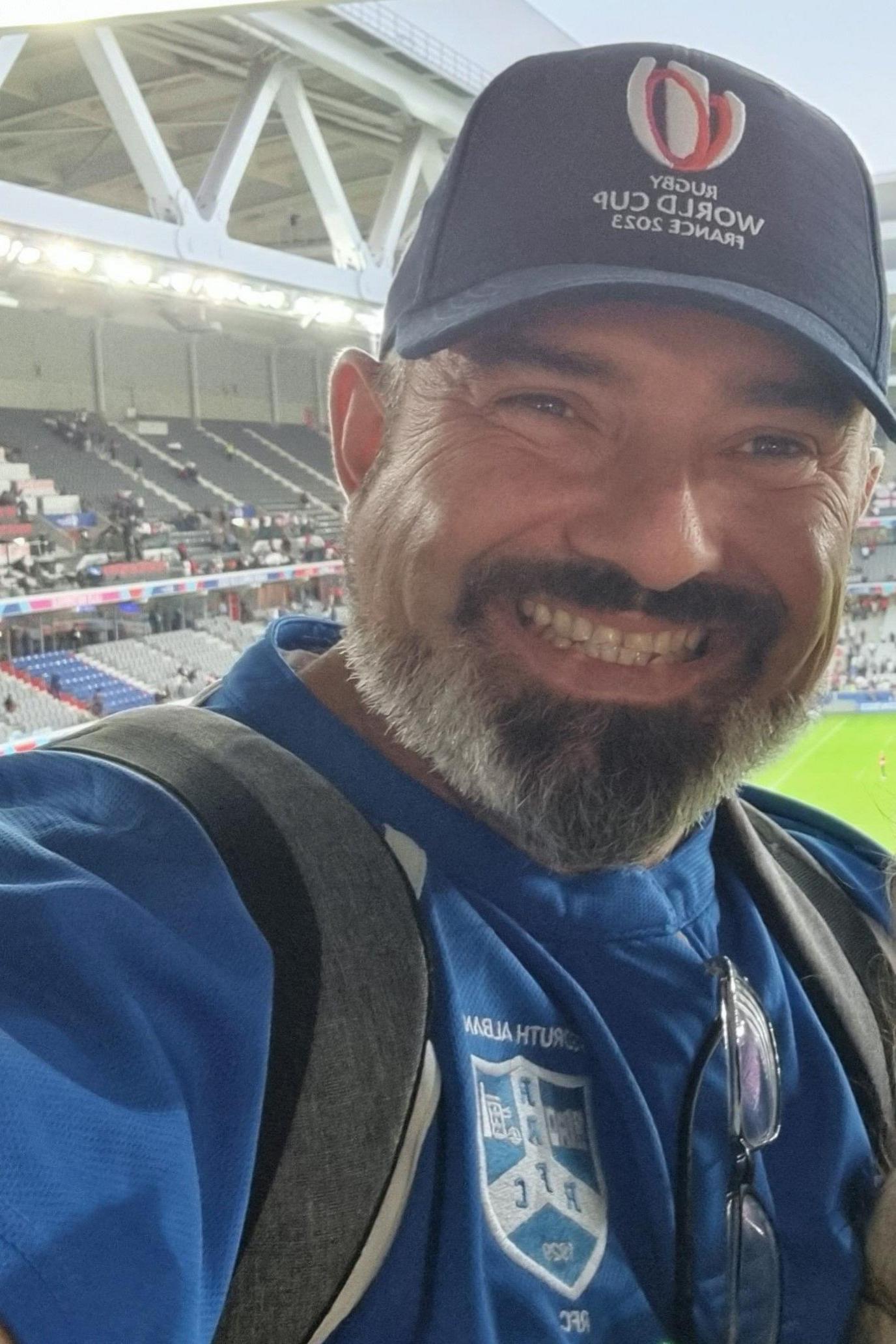 A selfie of Simon Ferguson as he is in stands overlooking a rugby pitch. He is wearing a blue cap with a logo and WORLD CUP printed on it and a blue club shirt. Grey backpack straps are visible across his shoulders. He is grinning and has a dark and grey beard and brown eyes.