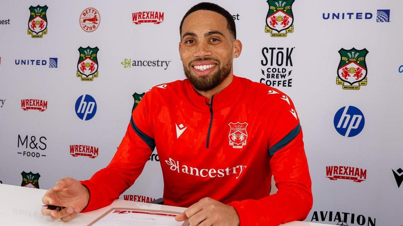 Zak Vyner sat in Wrexham club training gear with a pen 
