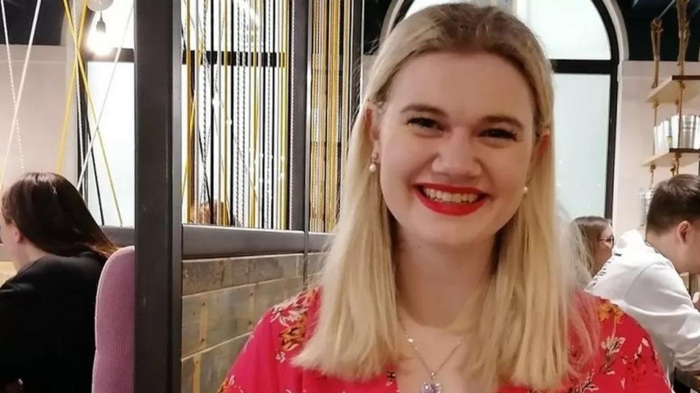 A woman wearing a red dress with a floral pattern smiles for the camera, she appears to be sat in a restaurant.