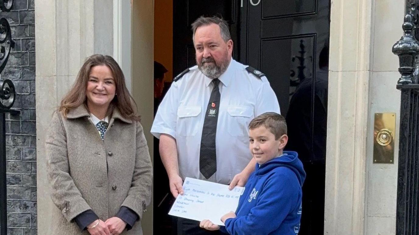 Roman and Jenny Riddell-Carpenter stand on the steps of 10 Downing Street. A security guard stands in between them in front of the door which is slightly open. The guard wears a white shirt with a black tie. He holds Roman's letter with him as they smile while looking away from the camera.