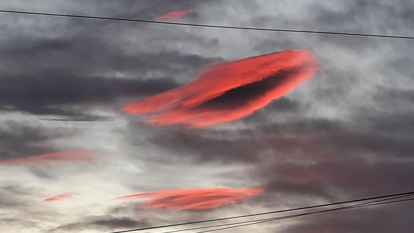 The cloud glows an orange-pink above industrial buildings that are behind a tall wire fence.