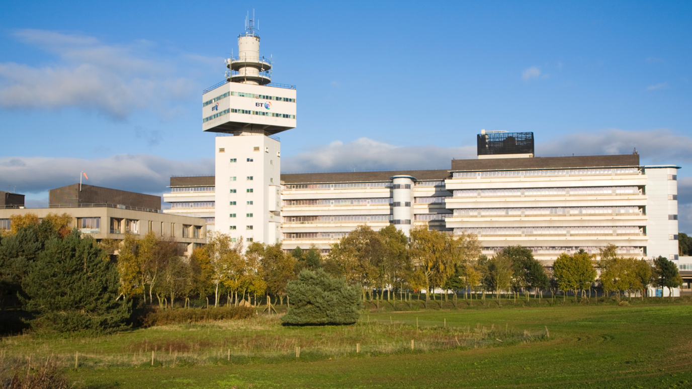 A big building spans across the whole image. On the left there is a tower, which has a mast on the top, and a sign which says BT. The building is white, and has lots of smalls windows going across it. 