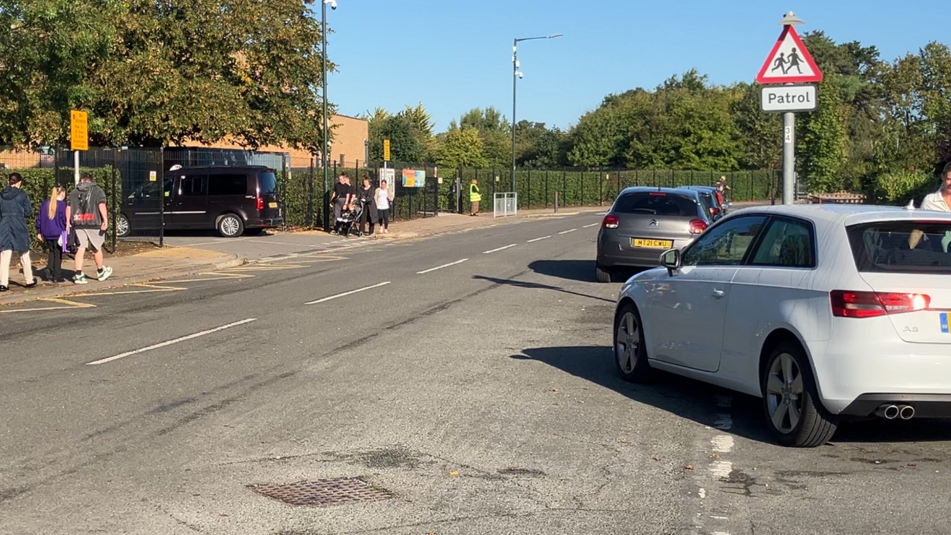 People walk by the side of a road towards a school entrance including a woman with a pushchair. A taxi can be seen at the school gates under a large green tree. On the left, yellow zig-zag lines are marked on the road and white cameras are attached to posts. To the right, a number of cars are parked on the roadside beneath a red, white and black "patrol" sign. More trees can be seen in the background under a blue sky.