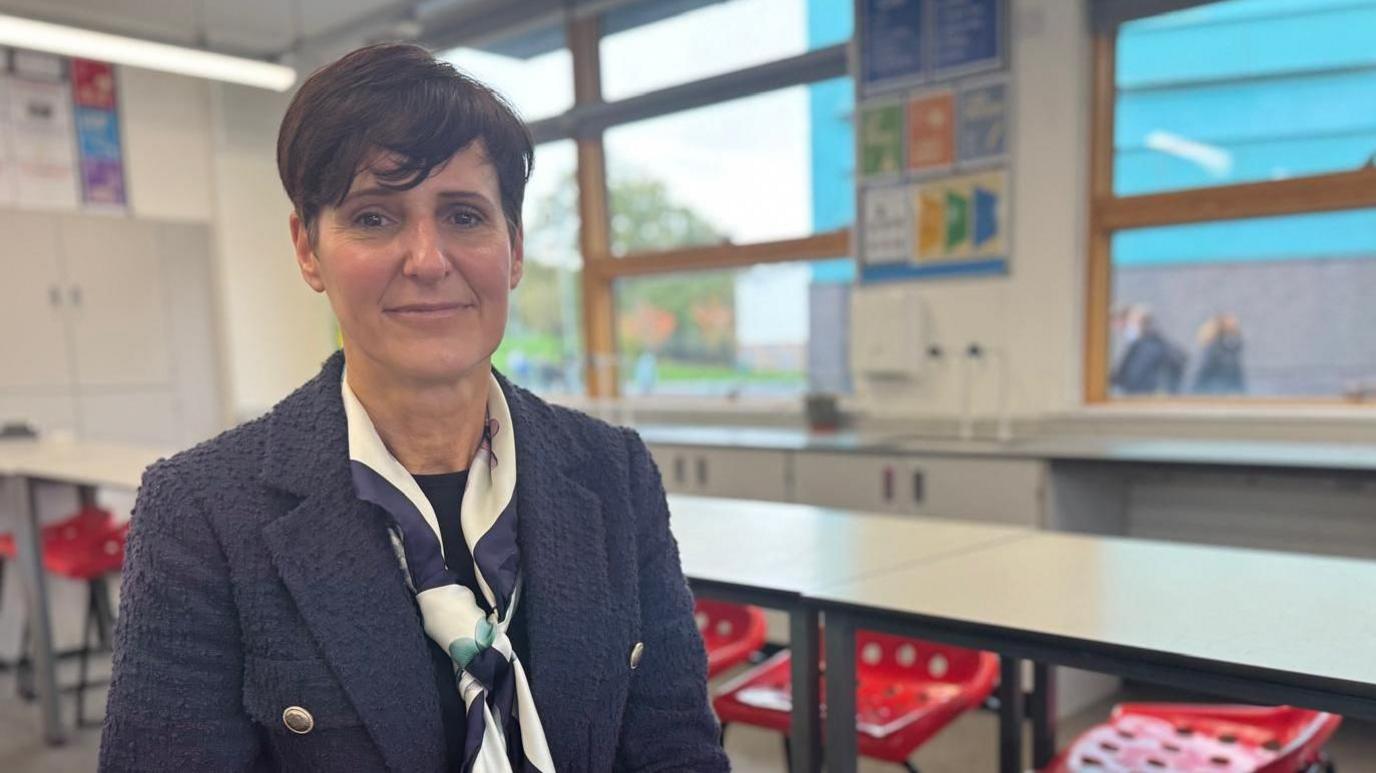 Becky Francis is seated at a table in a classroom wearing a dark textured jacket and a patterned scarf. The room has white walls, large windows letting in natural light, and posters with educational content on the wall. There are red plastic chairs with holes in the seat arranged around white tables.