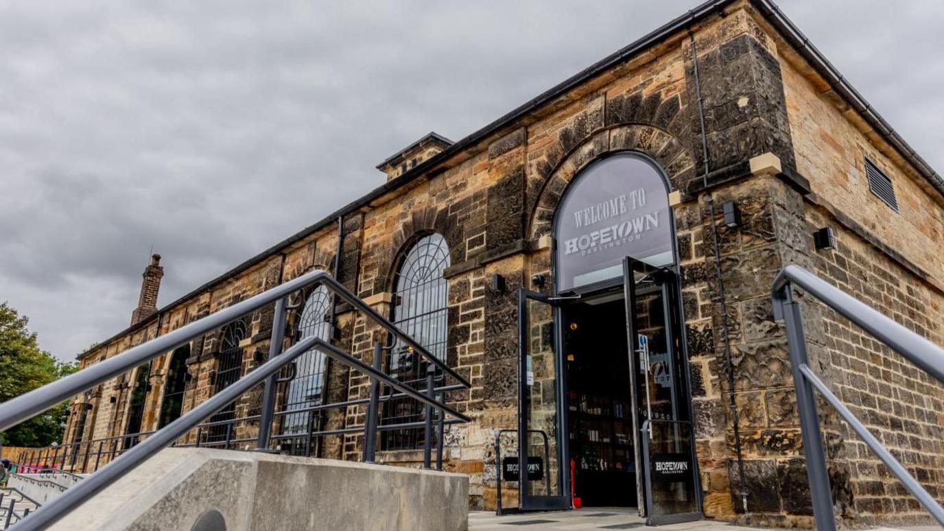The entrance to Hopetown, a single-storey dark brick building with signage, pictured from the foot of steps leading up to its front entrance There are large, arched windows with black leading.