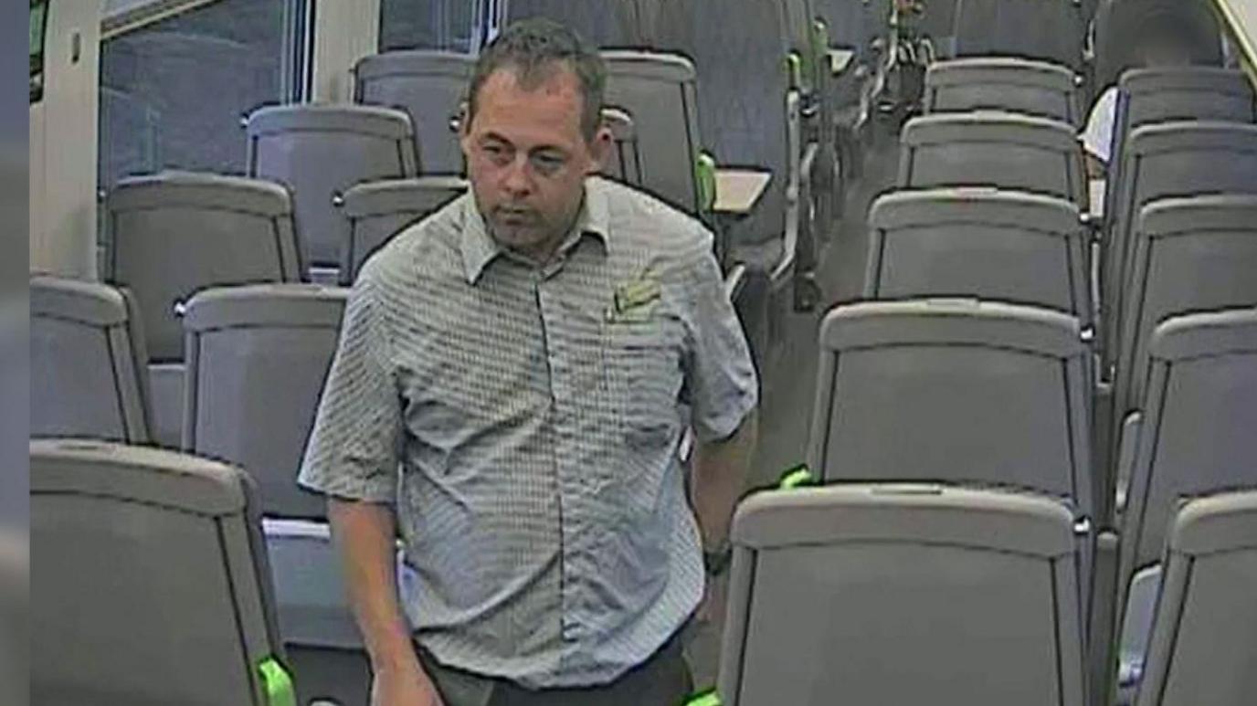 An image of a train conductor with brown hair and a green and white squared shirt on walking through a train carriage full of empty seats