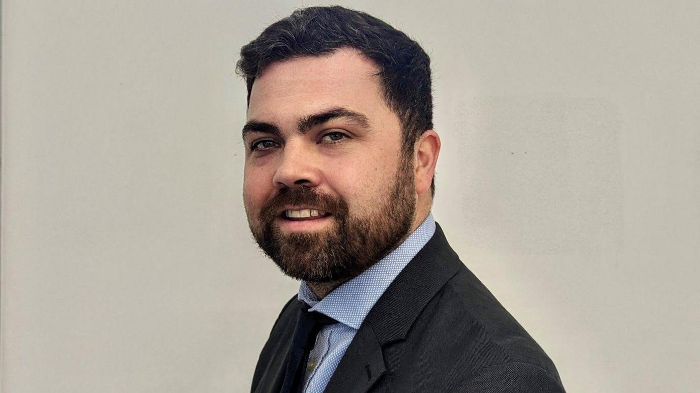 A man with dark hair and dark beard wears a dark suit with a blue shirt against a grey background. He is looking side on at the camera.