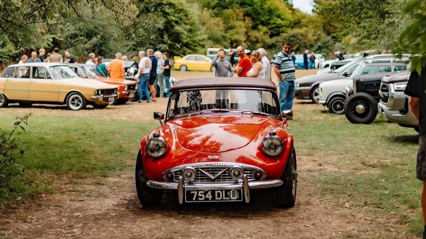 A red vintage car is parked on grass. People are surrounding it and taking photos. In the background are more parked vintage cars in varying colours.