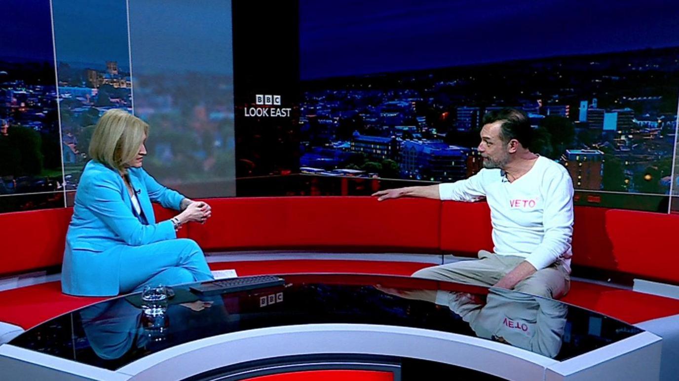 Mr Bone sits on a red sofa inside the BBC Look East studio. A reporter wearing a light blue suit sits opposite him during the interview.