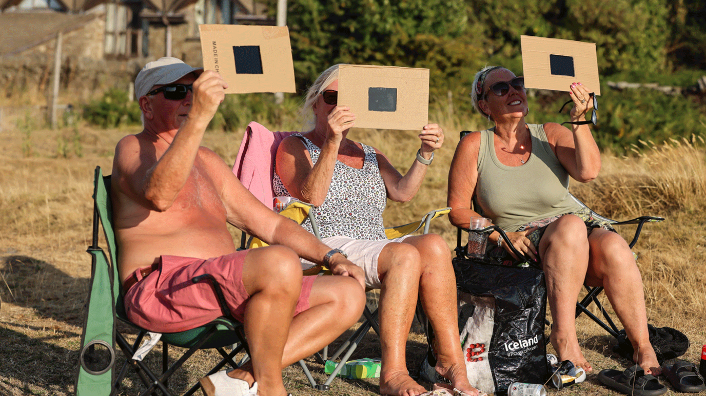 Three people sitting on camping chairs hold what looks like pieces of black glass in frames of cardboard to the sky to watch the partial eclipse