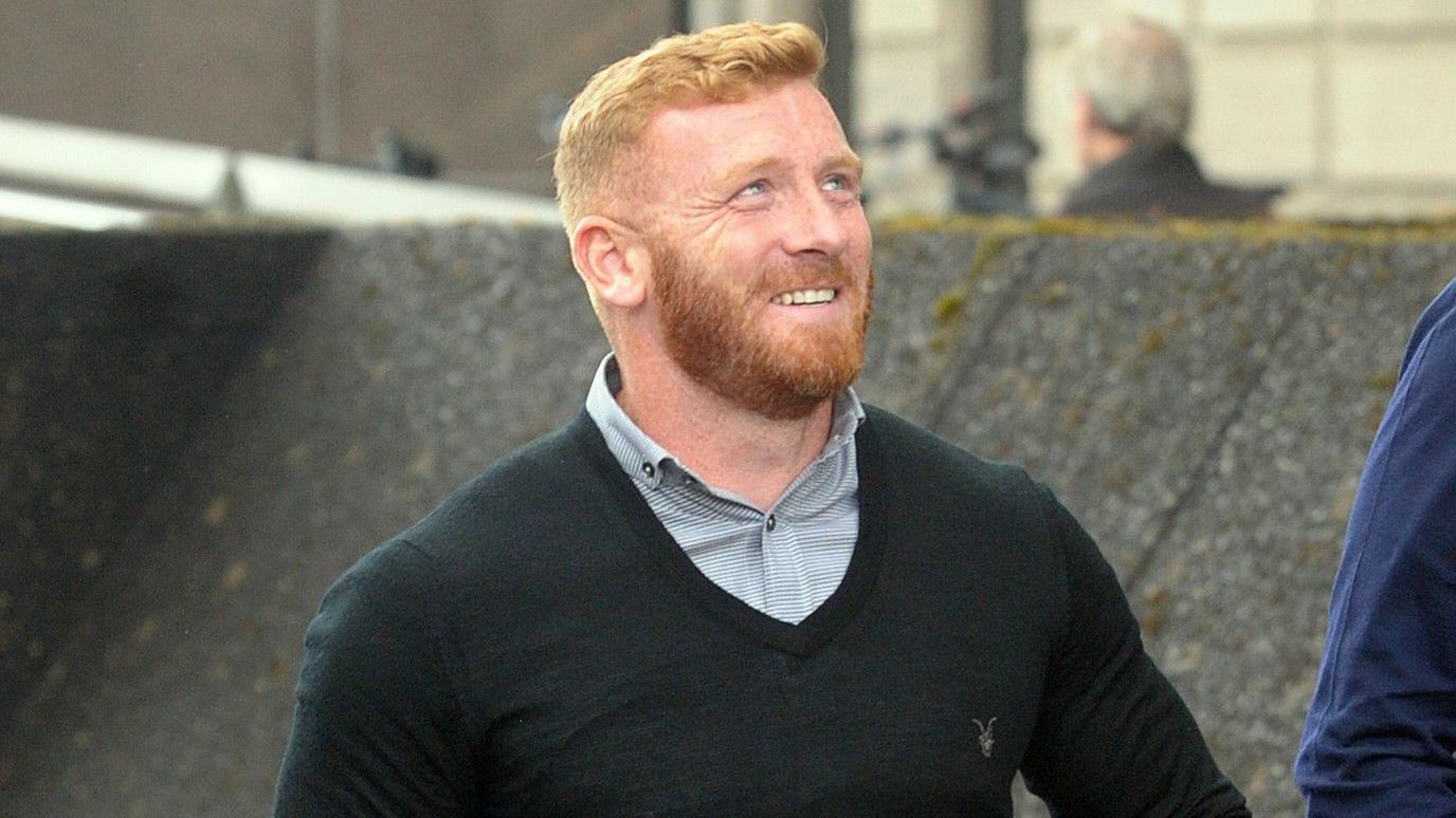 Head and shoulders shot of Ross Monaghan walking towards a court building. He is wearing a black v-neck jumper under a grey checked shirt. He has red hair and a well-trimmed beard. He is looking up to his left and grinning.