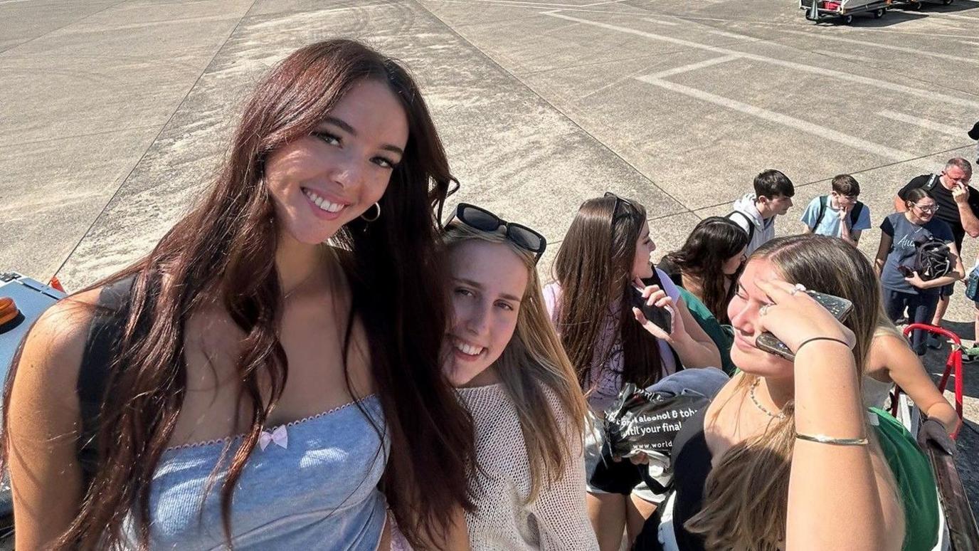 A girl with brown hair standing beside her two friends smiling at the camera