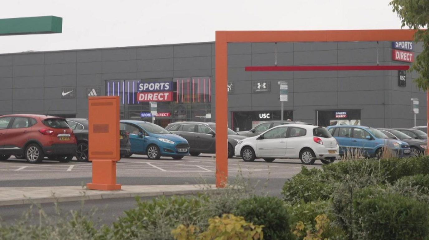 A sports direct store in a retail park. It is a large, cuboid, grey building. The logo for the shop is blue and red. The image is taken from a distance away in the car park, showing lots of parked vehicles near the storefront.