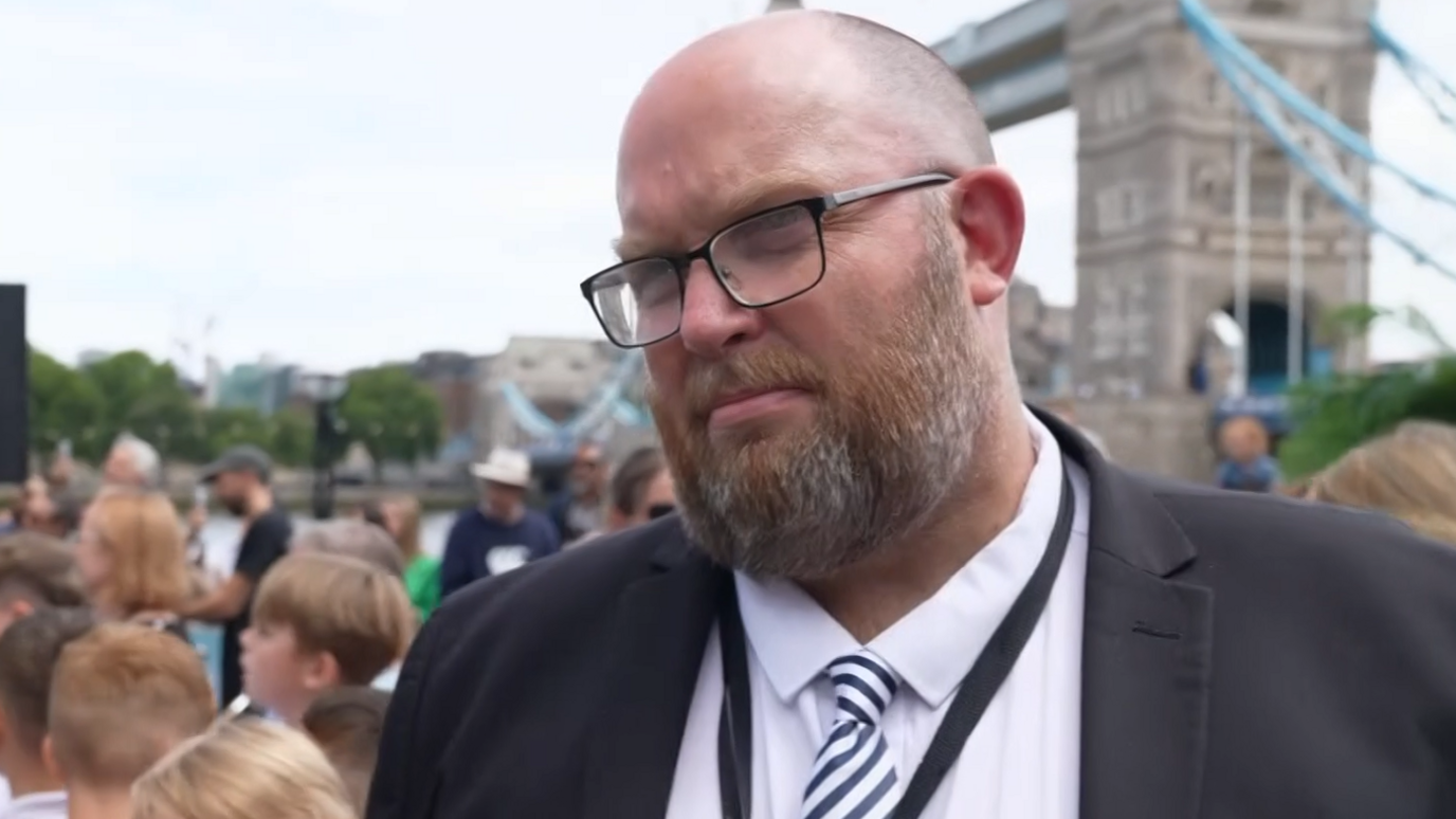 A head and shoulders picture of a man wearing glasses and a suit with a black blazer over a white shirt with a striped tie. Tower Bridge is in the background.