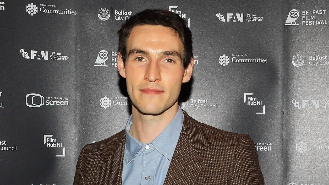 Éanna Hardwicke looking directly at the camera, he has dark hair and is wearing a brown suit with a light blue shirt.