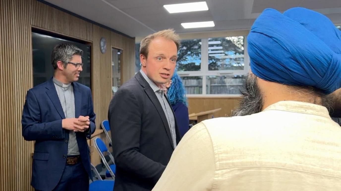 A group of men are stood talking in a room. The man in the foreground is wearing a turban and the man facing him is wearing a black jacket. There are other people in the background.