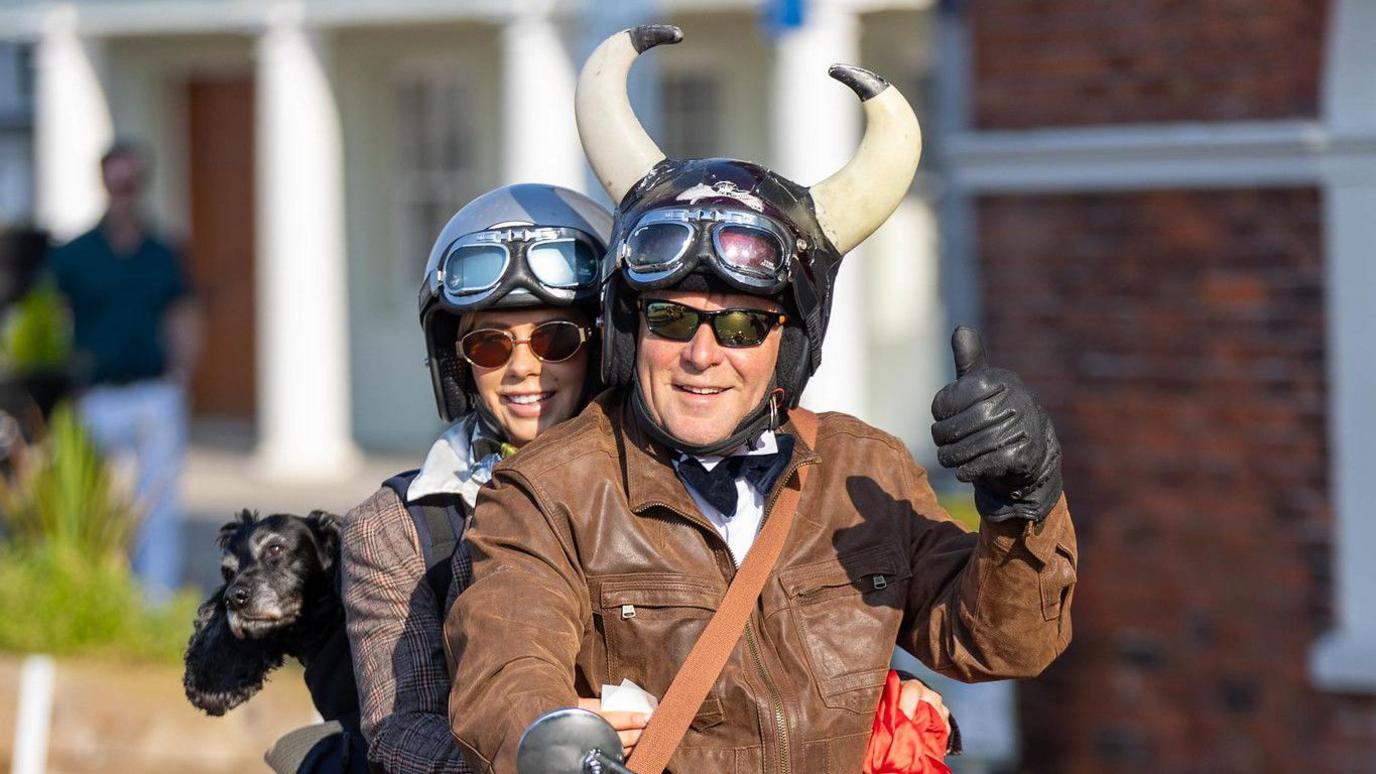 A man, woman and dog on a motorbike. The man is driving and the dog is peering from behind them both. The man is wearing a helmet with large horns - one on each side.