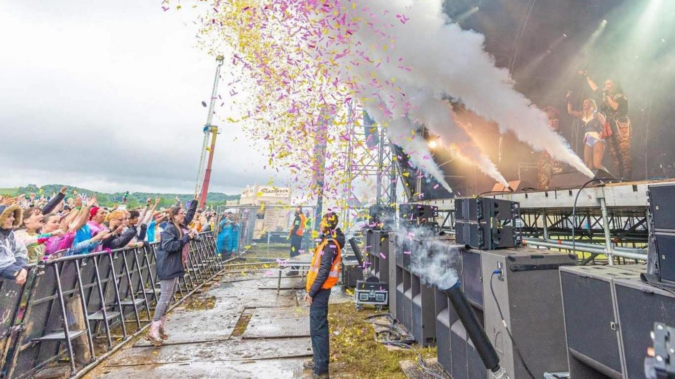 Kubix festival's stage photographed from the side. A security guard is standing in front of it as pink and yellow confetti flies in the air with stage smoke going off. Fans are lined behind a metal barrier and have their hands up in the air.