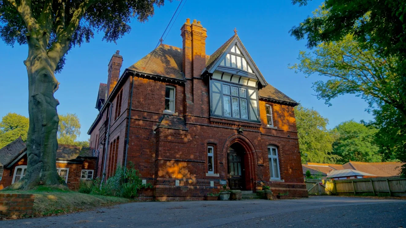 Brick building with Tudor frontage around first floor window. There are a further four windows in the front and a door. There are trees surrounding the property and a tarmac drive in front.