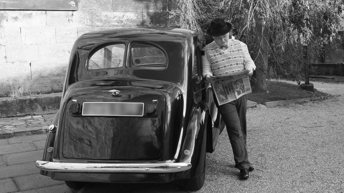 Black and white photo of Callum outside his car, reading a newspaper