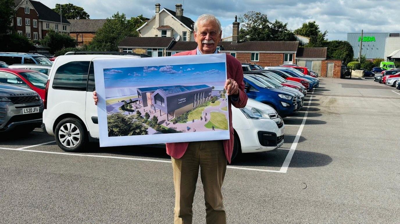 Councillor Ian Blair-Pilling stands in a car park while holding a large print of the artist's illustration of the new leisure centre. Mr Blair-Pilling has white hair and a moustache. He wears a red blazer jacket and light-brown coloured cargo trousers. There are cars and houses in the background.