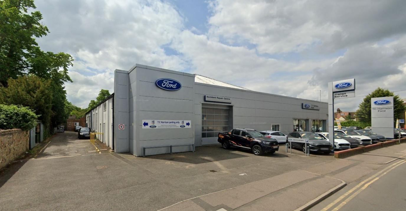 A Ford car garage. There are cars on the forecourt and large blue Ford signs on the grey building.