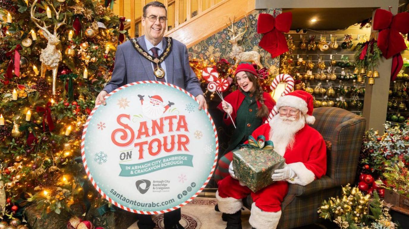 Lord Mayor Stephen Moutray, who is wearing a large gold, mayoral chain, stands holding a large, circular 'Santa on Tour' sign. He is stood beside Santa, who is sitting in a tartan armchair and holding a wrapped present. Beside him is a woman dressed up as an elf. The three are stood in a decorated room, with a Christmas tree and other Christmas decorations in the background.