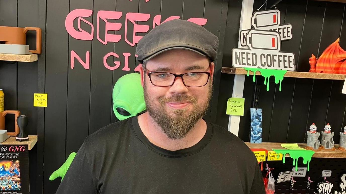 Michael, who is wearing a flat cap, and has a brown beard and glasses, stands behind the counter at his shop. Shelves with small figurines can be seen behind him.