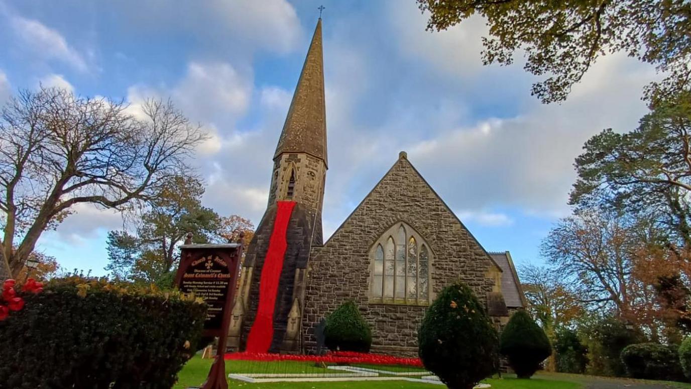 A church with hundreds of poppies hanging from the bell tower. A cross is on the lawn. Poppies are on bushes.
