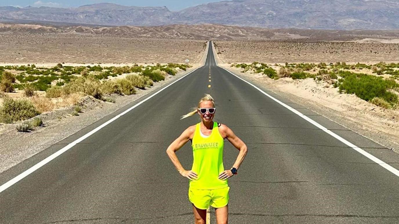 A smiling Laura Watts wearing a fluorescent yellow vest and shorts and sunglasses, posing on a long straight road across Death Valley with the road disappearing into the distance behind her.