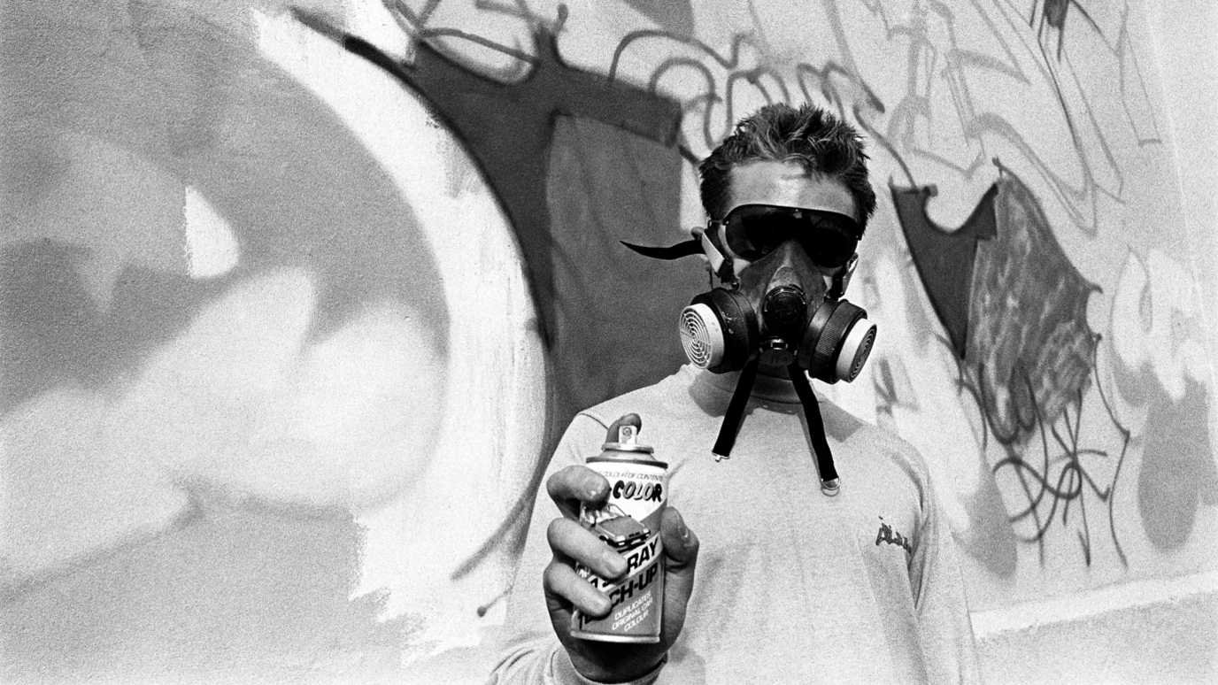 Picture taken by Beezer, of a man in a mask doing 3D graffiti art spray in St Paul's, Bristol, in 1985. The man is holding the spray can towards the camera with the graffiti wall behind him.