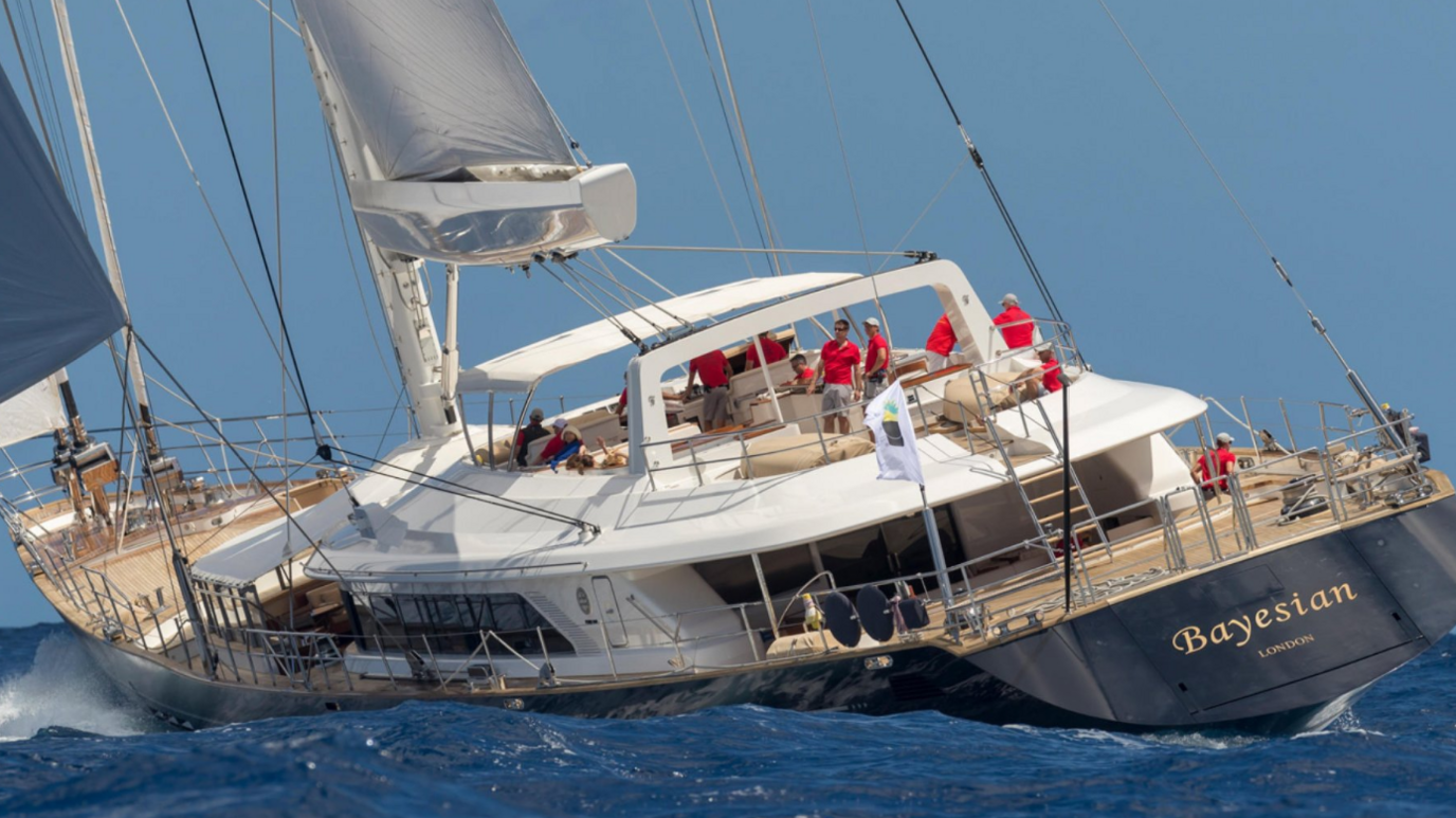 A photo of the Bayesian yacht pictured sailing. Several crew members in red tops can be seen. The ship's hull is a navy blue colour while it has one white sail and one blue sail.