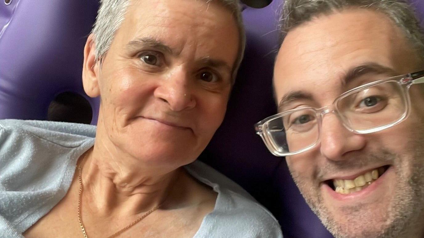 Michael Wheeler wearing clear framed rectangular glasses and smiling at the camera. Beside him is his mum, wearing a grey blouse and a gold necklace. She has dark eyes and very short grey hair. 