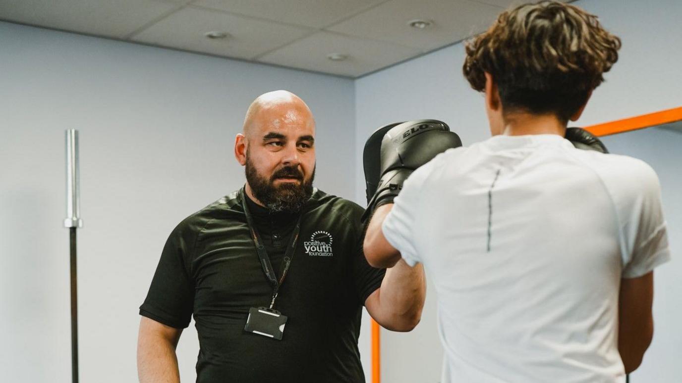 A man with a beard and a black top is facing the camera and boxing with a young male with dark hair and a white T-shirt with his back to the camera.