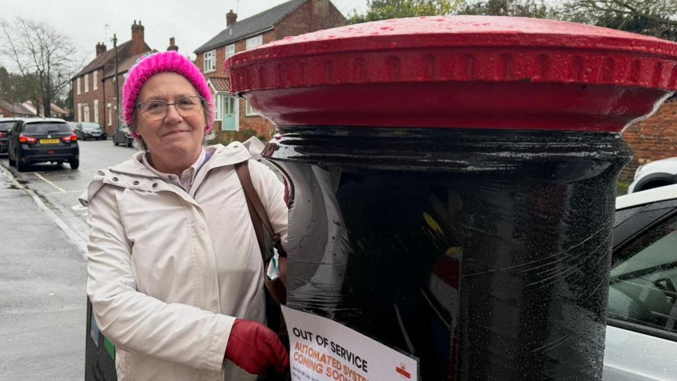 The photo shows a woman standing to the left of a postbox. She has glases on, and is wearing a cream coloured raincoat, bright red leather gloves, and a bright pink bobble hat. She is touching a postbox, which is to the right-hand side of the photo. It's covered in black wrapping. There is a sign that says 'Out of service. Automated system coming soon.' She's standing on a path, and there are cars in the background. 