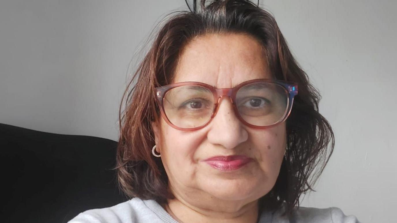 Samina has shoulder length brown hair and is wearing a light grey top and small gold hoop earrings and brown tortoise shell framed glasses.