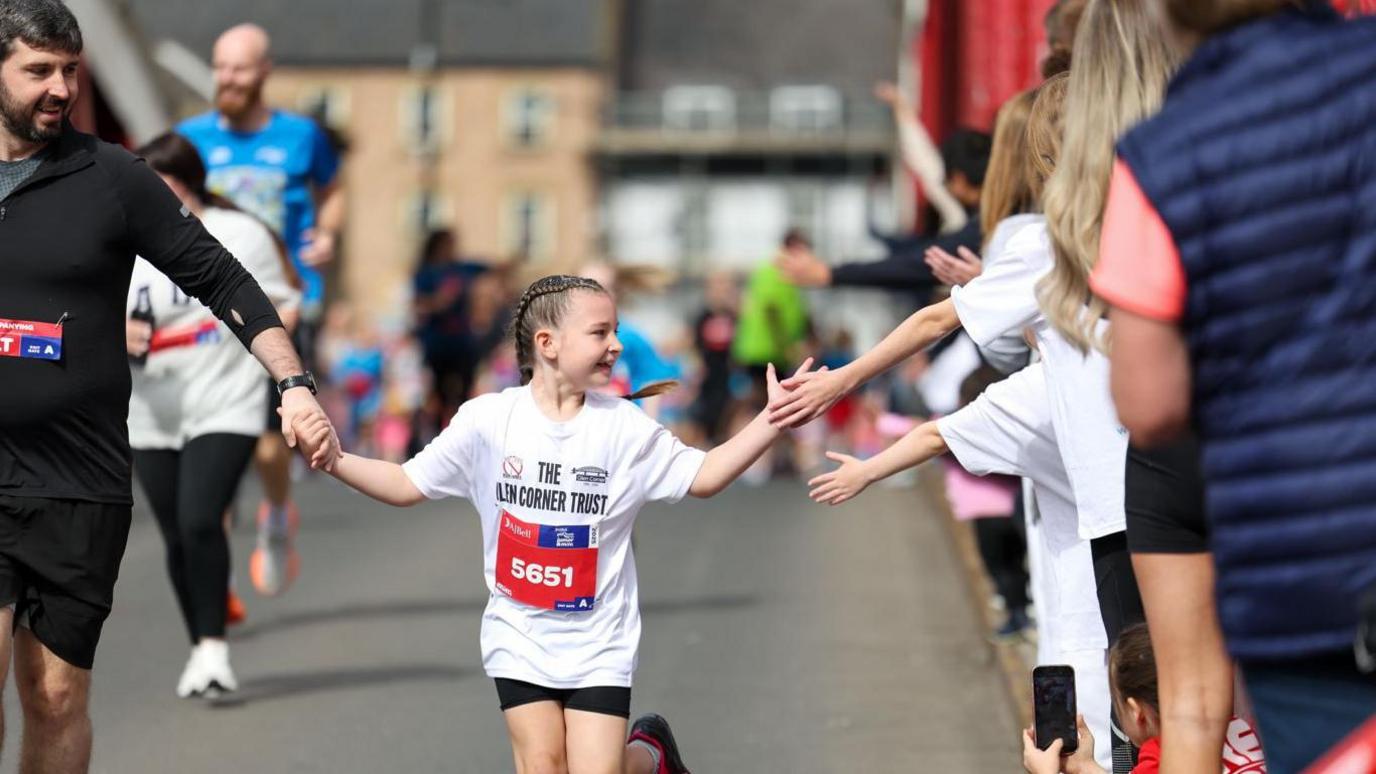 A young girl - aged about eight - taking part in a road race. She has braided brown hair and is wearing a white t-shirt with a race number in red. She is holding hands with an adult - a bearded man wearing a black t-shirt - and her other hand is reaching out and touching outstretched hand of people lining the street.