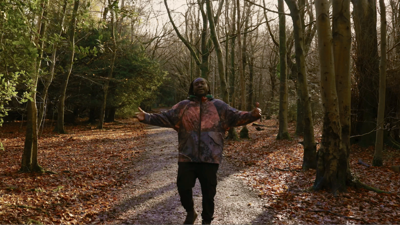 A man, centre, in a forest. He is wearing a burgundy and purple coat and black jeans. He is walking on a muddy path which has green trees either side and orange leaves on the floor.  