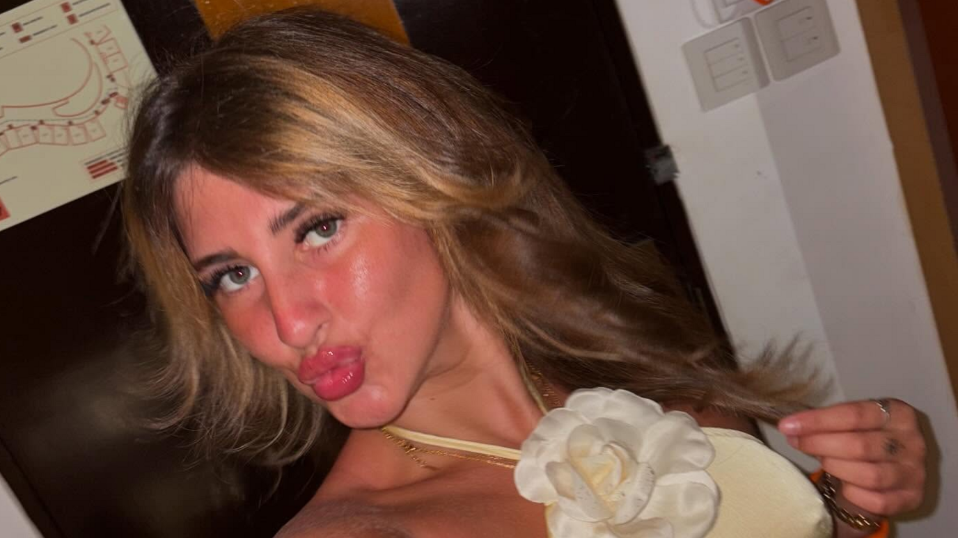 A selfie taken by Bella Culley of her wearing a yellow dress with a fabric white flower at her neck. She is pouting at the camera and holding a strand of hair in her other hand.
