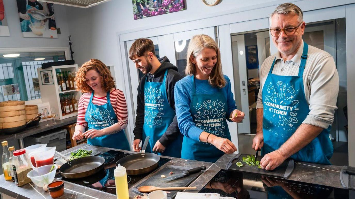 Two women and two men cooking using pans, saucepans and a chopping board.