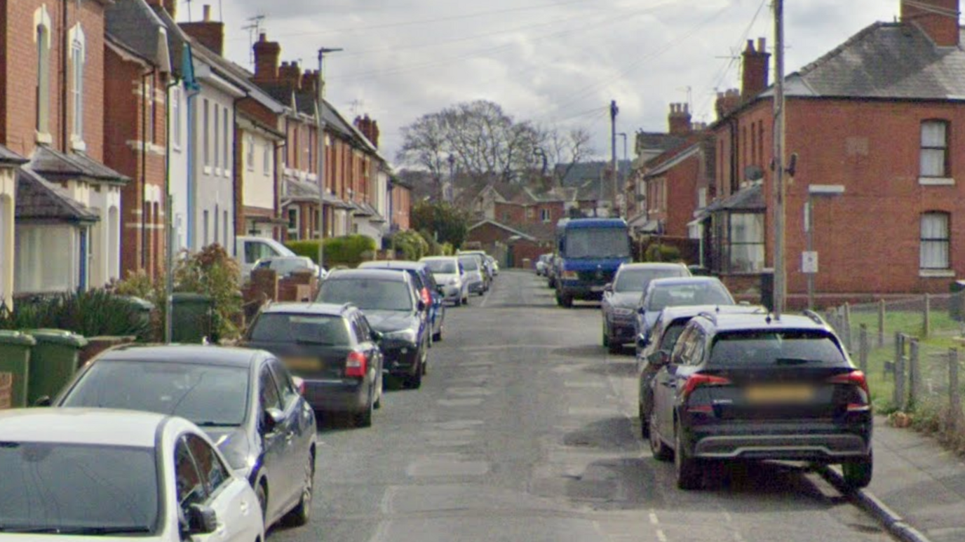 A residential street with terraced houses on either side and vehicles parked on both sides of the road