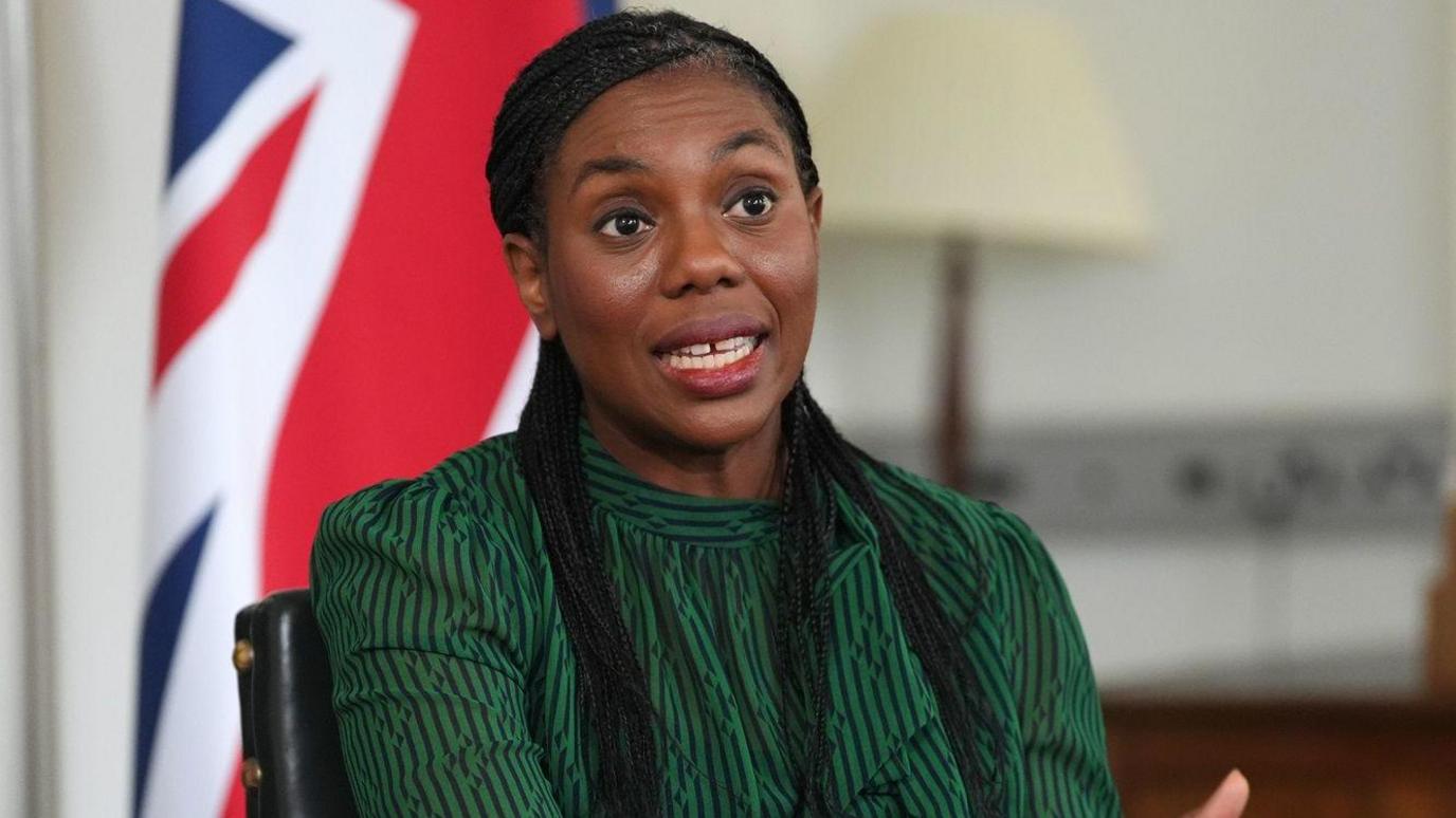 Kemi Badenoch talking and looking to the right of the camera. Her mouth is slightly open. She has long, dark braids and is sitting in front of a union jack flag wearing a green patterned top.