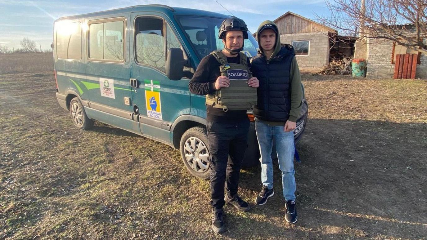 Vlad and his son Artem  stood in front of a green truck which has signs on it. Vlad is wearing a black jumper with a green army-style vest with black trousers. Artem is wearing a green jumper, a black gilet and blue trousers. The vehicle is on a grassy area with a house in the background.