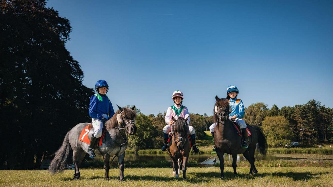 Three Shetland ponies with young riders in a grassy field with trees in the background