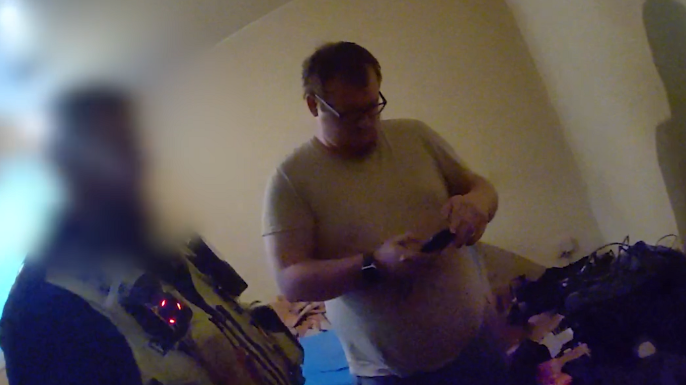 Matthew Webb being arrested by police in his home. He has short dark hair and is wearing a grey top, and holding his phone while police are waiting for him.
