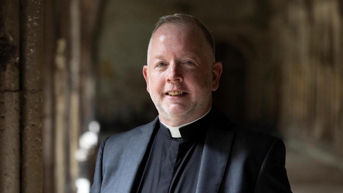 A man with short hair and stubble wearing a vicar's outfir, standing in a gothic-style passageway. He is looking at the camera and smiling.