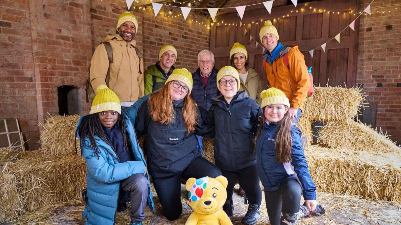 A group of people wearing hiking gear stand in a barn. A yellow pudsey bear sits in front. Stacks of hay can be seen behind them, with fairylights and bunting in the background.