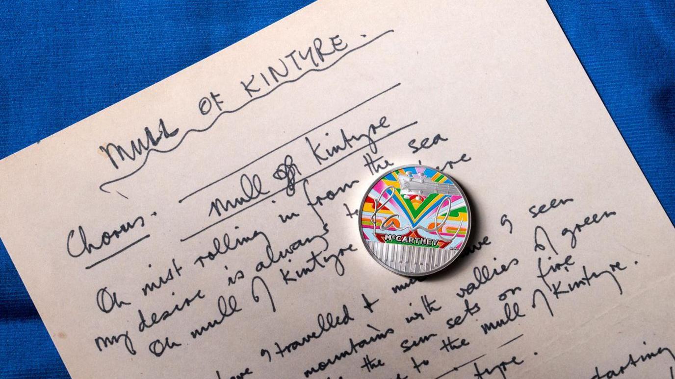 A silver coin with a rainbow design and Paul McCartney's signature rests on a copy of handwritten lyrics 