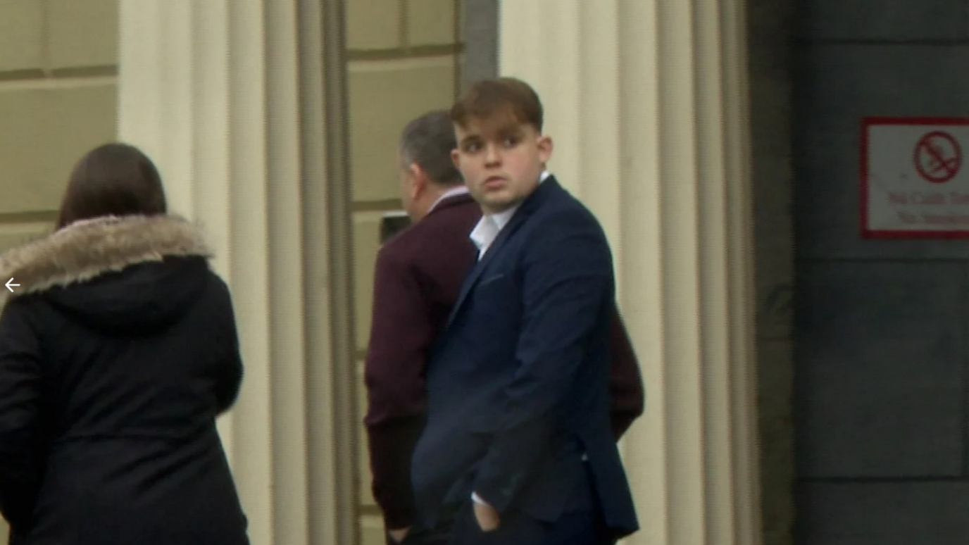 A man is wearing a suit and standing outside a building with large white pillars. A do not smoke sign is on the wall. A woman and a man are walking in the background.