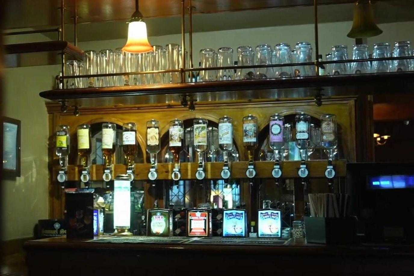 A dimly lit wooden bar inside Ye Olde Swiss Cottage pub, featuring a row of spirits in upside-down dispensers above beer taps. Glasses are stacked on a brass rack, and a vintage-style lamp is seen.